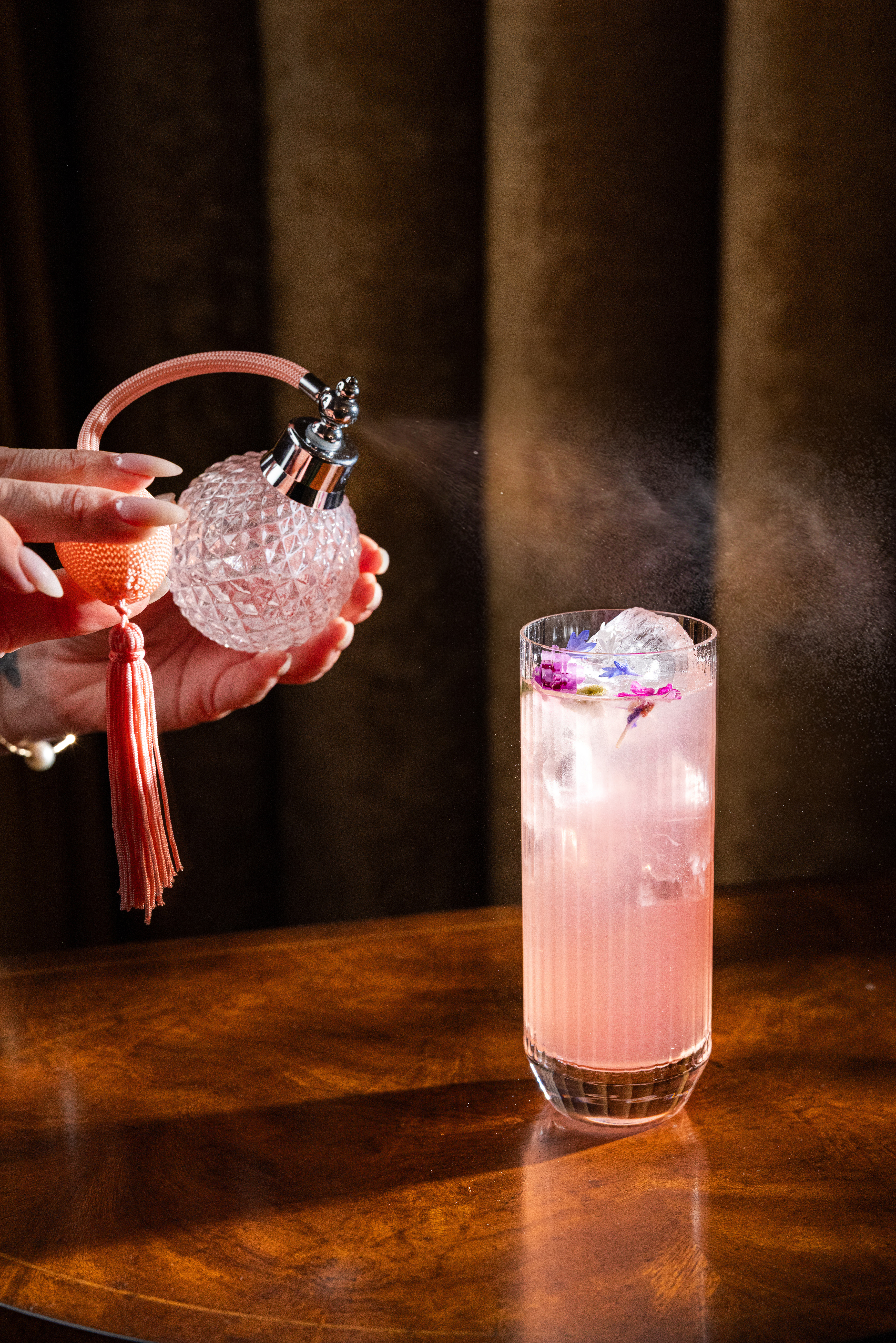 A signature cocktail being misted with a vintage crystal perfume atomizer, pink drink with edible flowers and ice