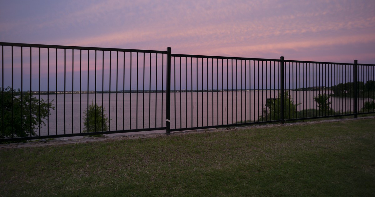 Lake Ray Hubbard at sunset seen through a black metal fence from a grassy Rockwall-area shoreline