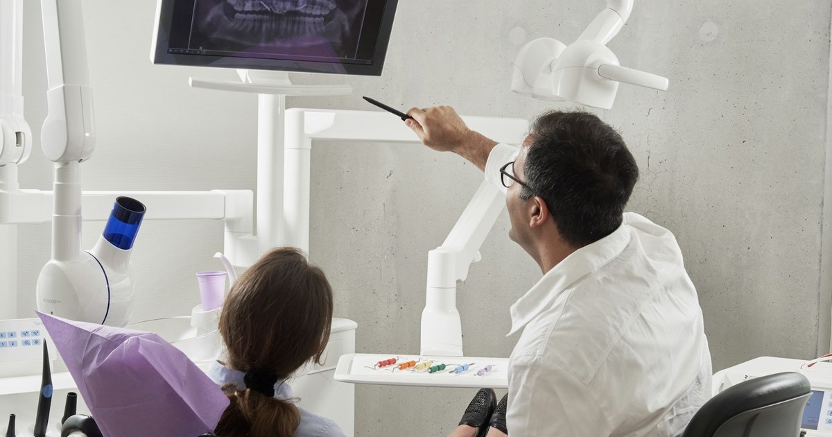 A dentist in a white coat pointing to a panoramic dental X-ray on a monitor for a patient seated in a modern dental chair